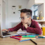 A glass wearing kid learning in his laptop.Laptop, juice, glass,pen,pencils,calculator on the table