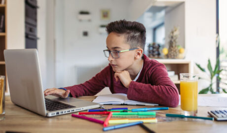 A glass wearing kid learning in his laptop.Laptop, juice, glass,pen,pencils,calculator on the table