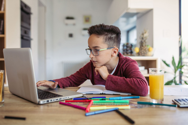 A glass wearing kid learning in his laptop.Laptop, juice, glass,pen,pencils,calculator on the table